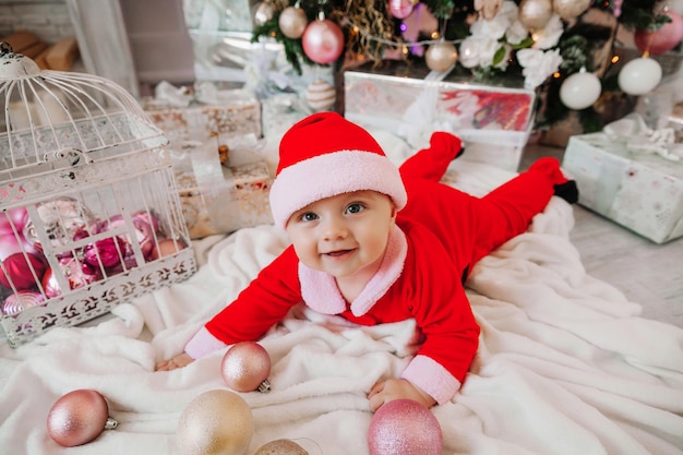 Ein süßes baby 6 monate alt in einem weihnachtsmannkostüm liegt auf einer decke in der nähe des weihnachtsbaums. weihnachten 2022