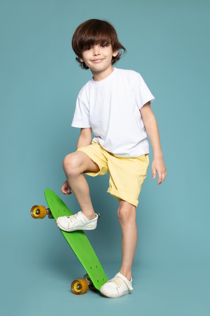 Ein süßes entzückendes süßes süßes Kind der Vorderansicht, das im weißen T-Shirt hält, das Skateboard auf dem blauen Raum hält