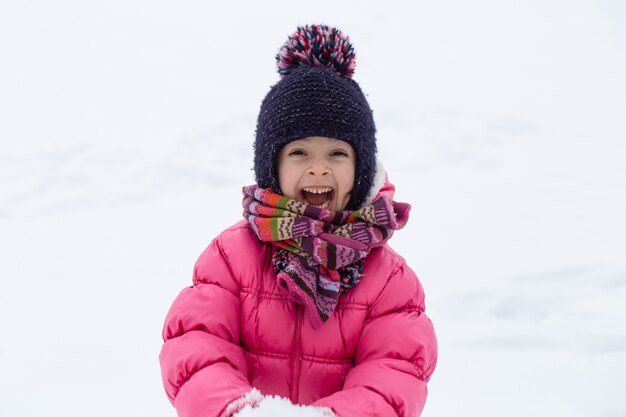 Ein süßes kleines Mädchen in einer rosa Jacke und einem Hut spielt im Schnee. Unterhaltungskonzept für Kinder im Winter.