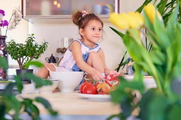 Ein süßes kleines Mädchen sitzt auf einem Tisch in einer Küche und versucht, Diätbrei zuzubereiten.