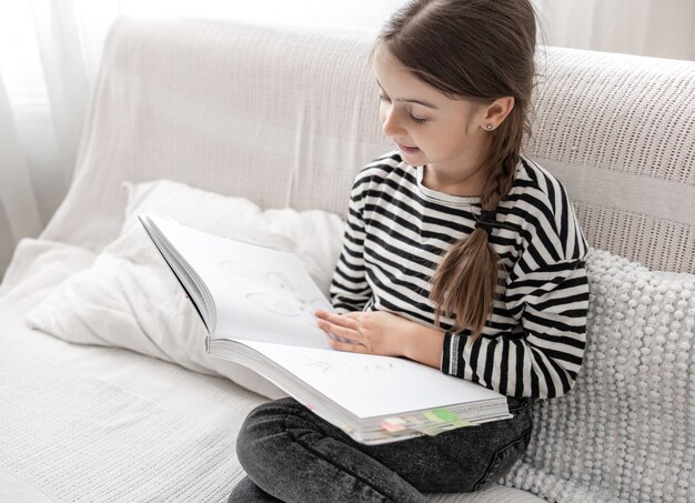Ein süßes kleines Mädchen untersucht begeistert ein Buch und sitzt zu Hause auf der Couch