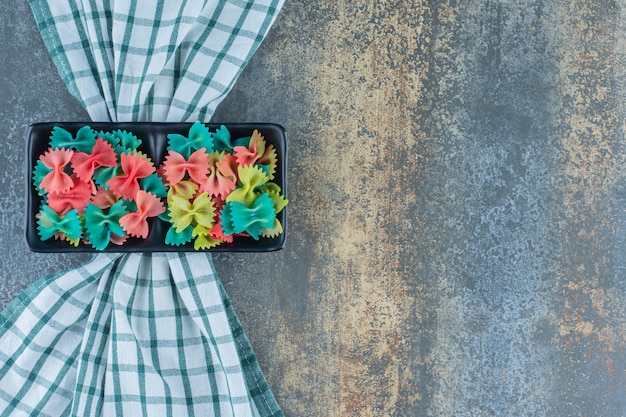 Ein Tablett mit bunten Farfalle-Nudeln auf dem Handtuch auf dem Marmorhintergrund.