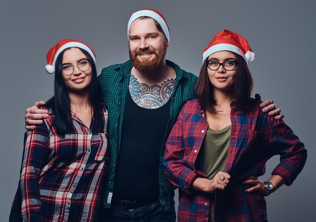 Ein tätowierter, bärtiger Mann und zwei brünette Frauen in Weihnachtskleidung posieren vor grauem Hintergrund.