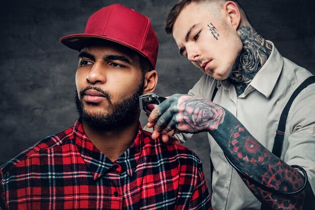 Ein tätowierter kaukasischer Mann in einem weißen Hemd, der den Bart von schwarzen Hipster-Männern in einem roten Fleece-Shirt schneidet.