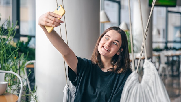 Ein Teenager-Mädchen sitzt in einer aufgehängten Hängematte und macht ein Selfie auf einem Smartphone