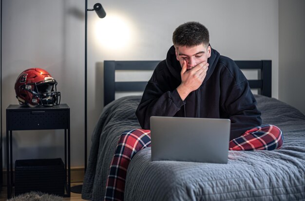 Ein Teenager sitzt in einem Zimmer auf einem Bett und benutzt einen Laptop