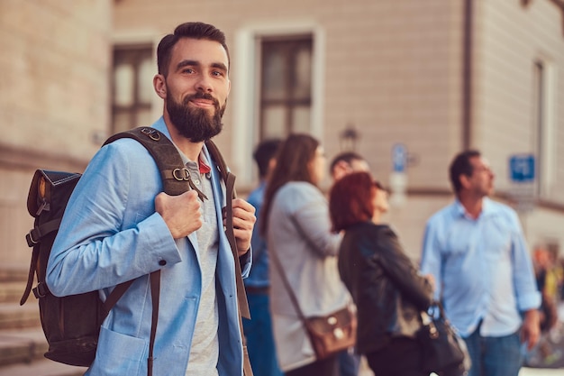 Ein Tourist mit Vollbart und Haarschnitt, der Freizeitkleidung trägt, hält während der Exkursion in Europa einen Rucksack in der Hand und schreibt eine SMS auf ein Smartphone, das auf einer antiken Straße steht.