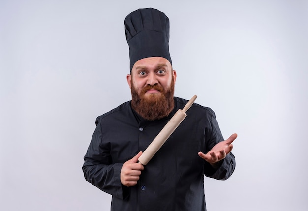 Ein überraschter bärtiger Kochmann in der schwarzen Uniform, der Nudelholz hält, während er Kamera mit dem Ausdruck betrachtet, weiß nicht, was auf einer weißen Wand zu tun ist