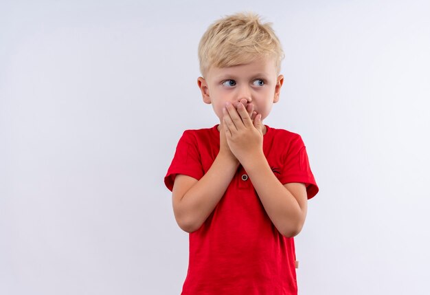 Ein überraschter kleiner süßer blonder Junge im roten T-Shirt, der Hände auf seinem Mund hält, während Seite auf einer weißen Wand schaut