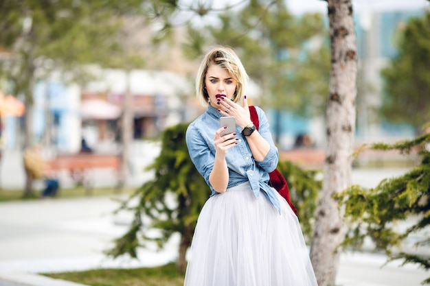 Ein überraschtes Mädchen mit kurzen blonden Haaren und leuchtend rosa Lippen, die ein Smartphone betrachten