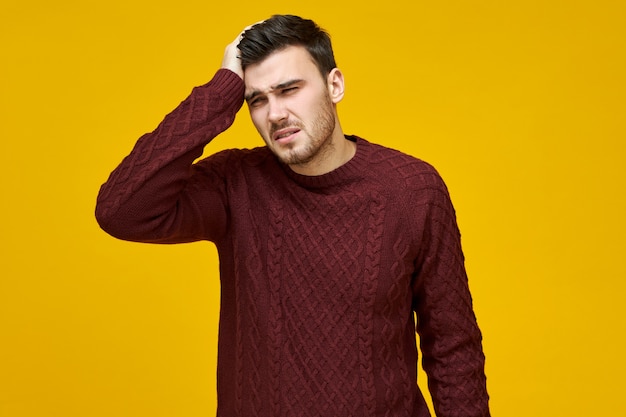 Ein unglücklicher junger Mann in einem warmen Pullover, der Erkältung, Grippe oder einen schlechten Arbeitstag hat und unter spaltenden Kopfschmerzen leidet