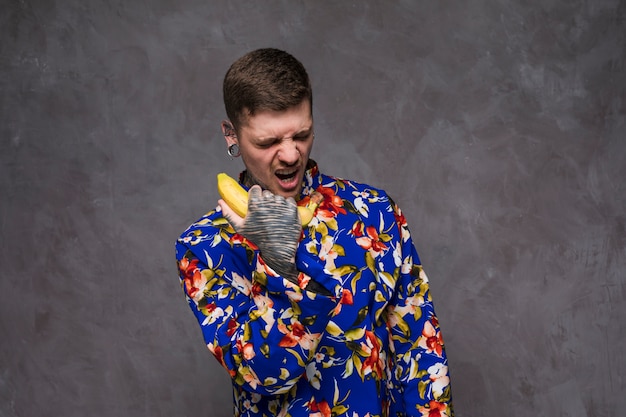 Ein verärgerter junger Mann mit tätowiert auf seiner Hand schreit am Bananentelefon gegen graue Wand