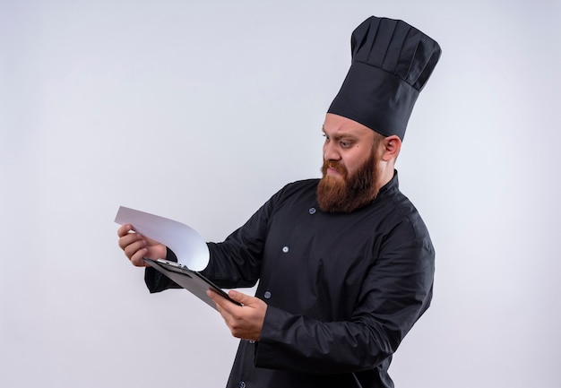 Ein verwirrter bärtiger Kochmann in der schwarzen Uniform, die einen leeren Ordner mit aggressivem Ausdruck auf einer weißen Wand betrachtet
