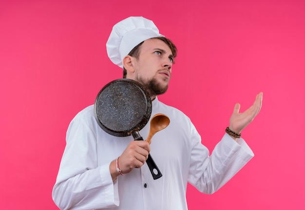 Ein verwirrter junger bärtiger Kochmann in der weißen Uniform, die Bratpfanne mit Holzlöffel auf einer rosa Wand hält