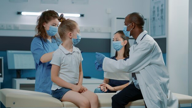 Ein vielfältiges medizinisches Team analysiert die Röntgenergebnisse mit Mutter und Kind im Kabinett. Hausarzt und Krankenschwester erklären Frau und kleinem Mädchen bei Termin die Röntgendiagnose.