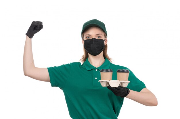 Ein weiblicher Kurier der Vorderansicht in den schwarzen Handschuhen der grünen Uniform und der schwarzen Maske, die Kaffeetassen hält