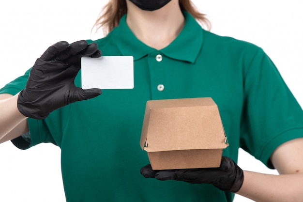 Ein weiblicher Kurier der Vorderansicht in den schwarzen Handschuhen der grünen Uniform und in der schwarzen Maske, die Lebensmittel-Lieferpaket und weiße Karte halten