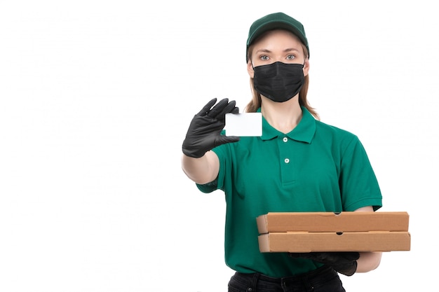 Ein weiblicher Kurier der Vorderansicht in den schwarzen Handschuhen der grünen Uniform und in der schwarzen Maske, die Nahrungsmittellieferboxen hält