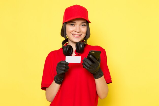 Ein weiblicher Kurier der Vorderansicht in den schwarzen Handschuhen der roten Uniform und der roten Kappe, die weiße Karte und Telefon mit Lächeln hält