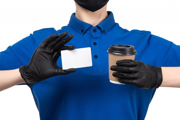 Ein weiblicher Kurier der Vorderansicht in der blauen einheitlichen schwarzen Maske und in den Handschuhen, die Kaffeetasse und weiße Karte halten