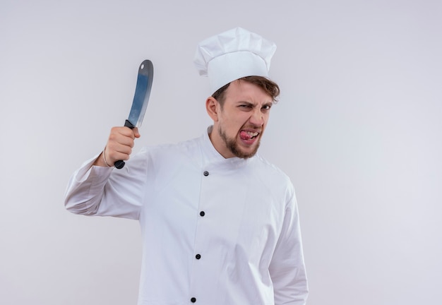 Ein wütender junger bärtiger Kochmann in der weißen Uniform, die Fleischbeil hält, während auf einer weißen Wand schaut