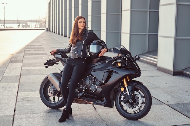 Ein wunderschönes Biker-Mädchen mit schwarzer Lederjacke, das sich vor einem Gebäude auf ihr Superbike stützt.