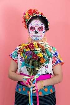 Eine als catrina verkleidete junge frau in mexikanischem kostüm, die mit schlichtem hintergrund und blumen posiert