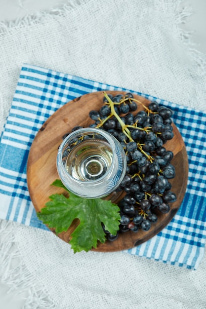 Eine Ansammlung schwarzer Trauben mit Blatt und einem Glas Wein auf weißer Oberfläche mit blauer Tischdecke