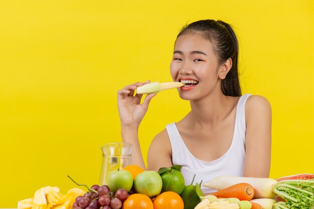 Eine asiatische Frau, die ein weißes Trägershirt trägt. Baby Mais essen gehen und auf dem Tisch gibt es viele Früchte.