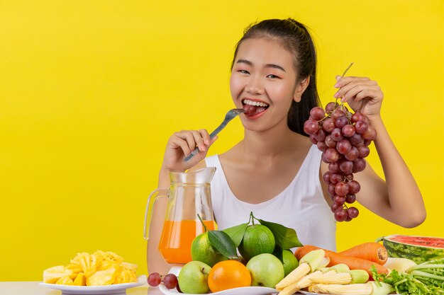 Eine asiatische Frau, die ein weißes Trägershirt trägt. Die linke Hand hält eine Weintraube. Die rechte Hand nimmt die Trauben zum Essen und der Tisch ist voll mit verschiedenen Früchten.