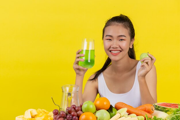 Eine asiatische Frau, die ein weißes Trägershirt trägt. Mit der rechten Hand ein Glas Apfelsaft halten Mit der linken Hand einen Apfel halten Und der Tisch ist voll mit verschiedenen Obstsorten.