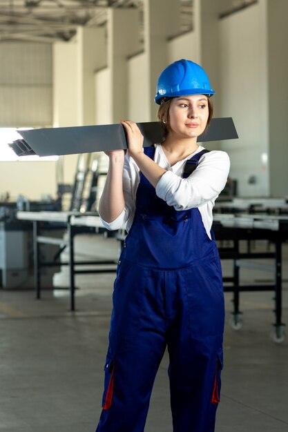 Eine attraktive junge Dame der Fernentfernungsansicht im blauen Bauanzug und im Helm, die schweres metallisches Detail während des Architekturbaus der Tagesgebäude halten