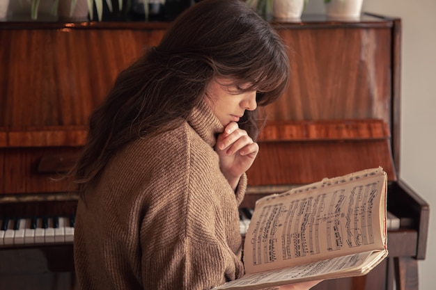 Eine attraktive junge frau in einem gemütlichen pullover sitzt neben dem klavier und schaut auf die noten.
