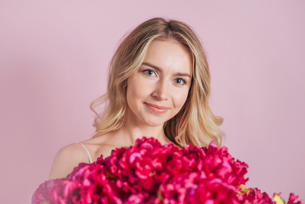 Eine attraktive lächelnde blonde junge Frau mit rotem Blumenblumenstrauß gegen rosa Hintergrund