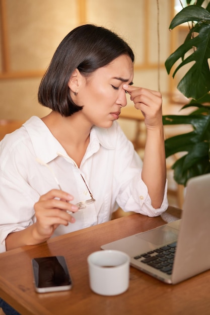 Eine berufstätige Frau fühlt sich müde, wenn sie sich die Augen reibt, nachdem sie einen Laptop benutzt hat, der müde ist, im Café mit Kaffee zu sitzen