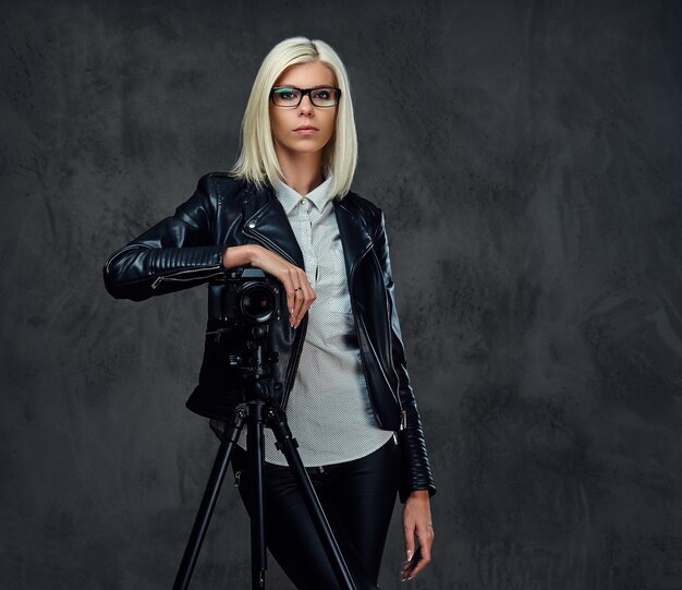 Eine blonde Fotografin in einer schwarzen Lederjacke hält eine professionelle Digitalkamera und ein Stativ.