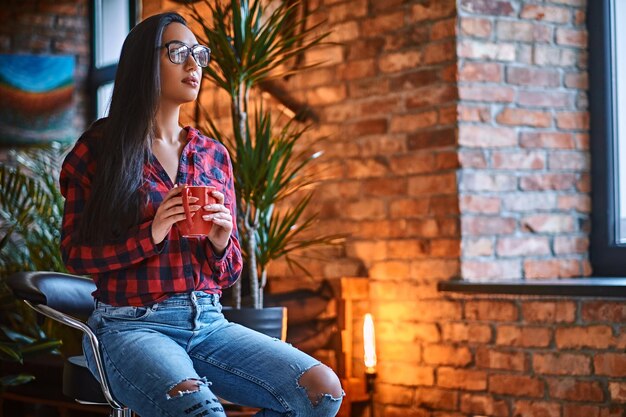 Eine brünette Hipster-Frau in einem Fleece-Shirt und Jeans trinkt Kaffee in einem Raum mit Loft-Interieur.