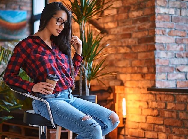 Eine brünette Hipster-Frau in einem Fleece-Shirt und Jeans trinkt Kaffee in einem Raum mit Loft-Interieur.