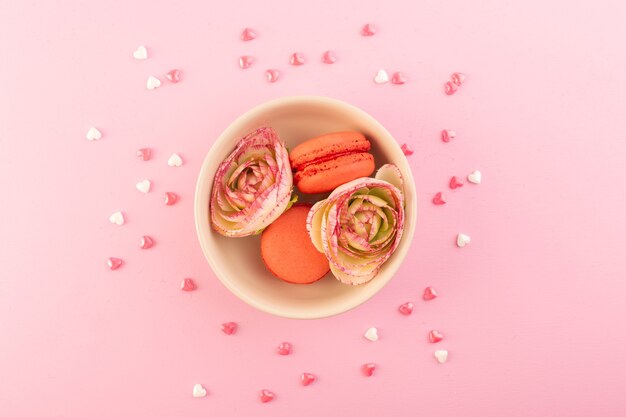 Eine bunte französische Macarons der Draufsicht mit Blumen auf dem rosa Schreibtischkuchen-Kekszuckersüß