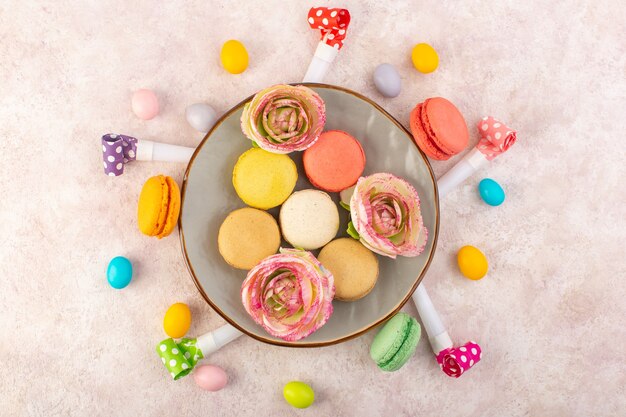 Eine bunte französische Macarons der Draufsicht mit Süßigkeiten und Blumen auf dem süßen Zucker der rosa Schreibtischkuchen