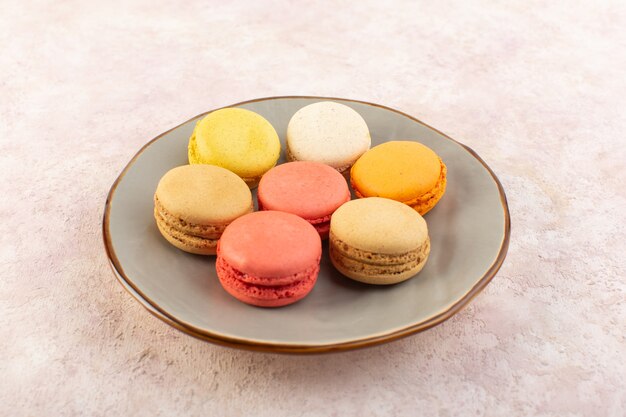 Eine bunte französische Macarons der Vorderansicht innerhalb der Platte auf dem rosa Schreibtisch