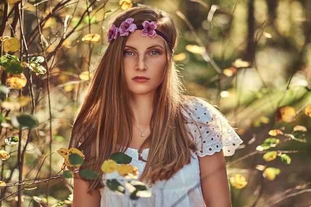 Eine charmante schöne Frau in einem weißen Kleid und einem lila Kranz auf dem Kopf, die in einem grünen Herbstwald posiert.