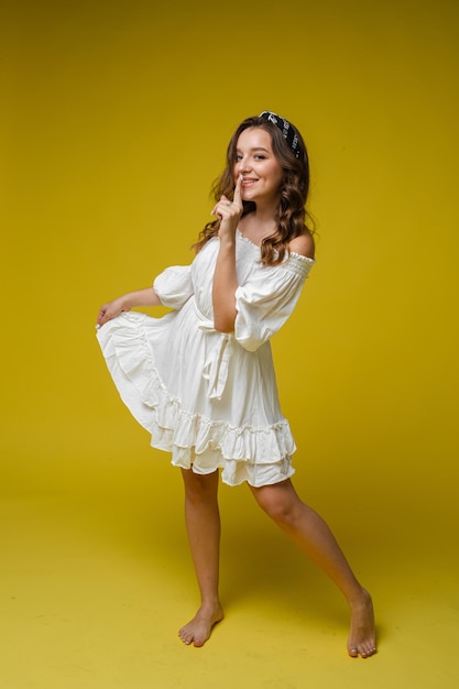 Eine coole, helle Frau in einem weißen Kleid mit schönem lockigem Haar und einem entzückenden Lächeln im Studio auf gelbem Hintergrund