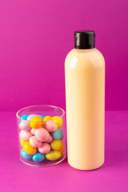 Eine cremefarbene Plastik-Shampoo-Dose mit Vorderansicht mit schwarzer Kappe und bunten Bonbons, die auf dem Schönheitshaar der lila Hintergrundkosmetik isoliert sind