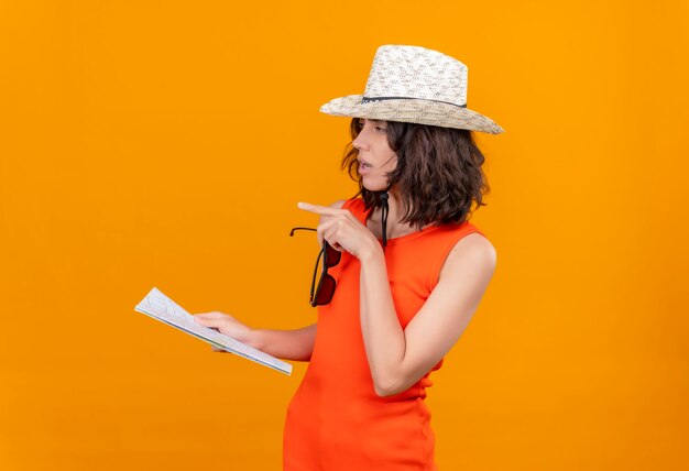 Eine denkende junge Frau mit kurzen Haaren in einem orangefarbenen Hemd, das Sonnenhut hält Karte mit Sonnenbrille hält