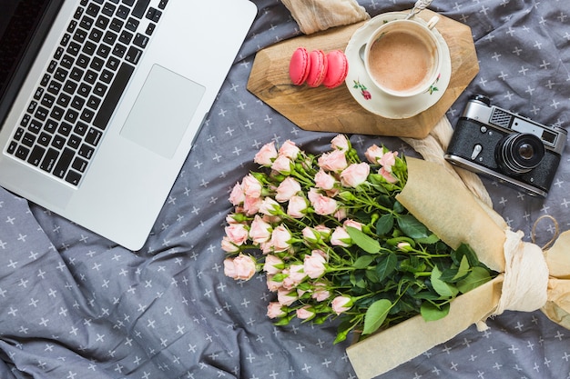 Eine draufsicht auf den laptop; makrone; kaffeetasse; kamera- und blumenstrauß auf grauer tischdecke