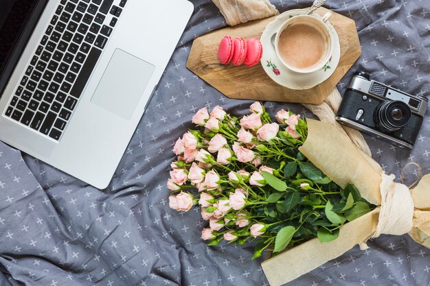 Eine Draufsicht auf den Laptop; Makrone; Kaffeetasse; Kamera- und Blumenstrauß auf grauer Tischdecke