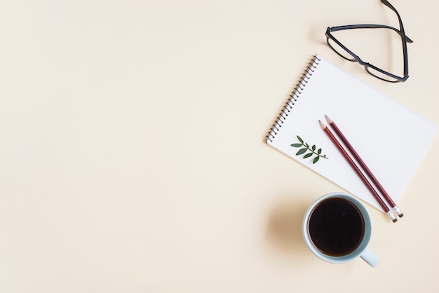 Eine Draufsicht der Teetasse; Brille und Bleistift auf Spiralblock vor beigem Hintergrund