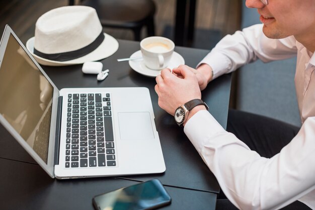 Eine Draufsicht eines jungen Mannes, der Laptop mit Kaffee betrachtet; Hut und Handy auf dem Tisch