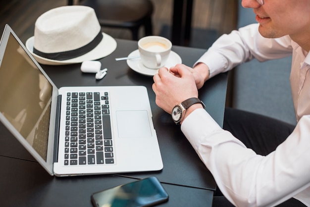 Eine Draufsicht eines jungen Mannes, der Laptop mit Kaffee betrachtet; Hut und Handy auf dem Tisch